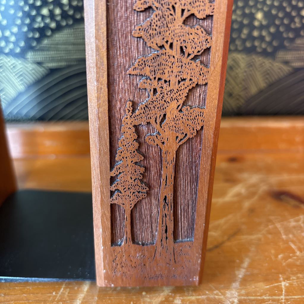 A wooden bookend with a laser cut image of a tall tree on the front sitting on a wooden surface in front of a black and gold wallpaper.