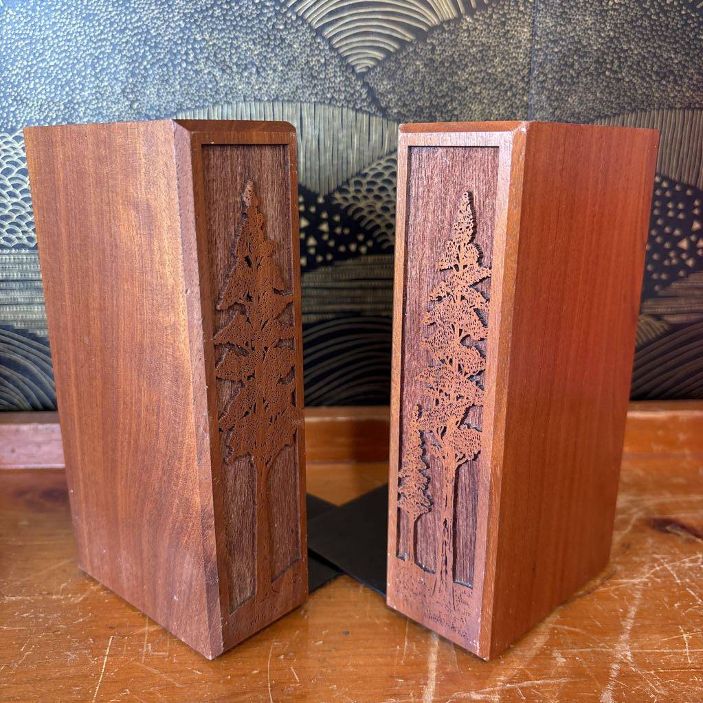 Two wooden bookends with laser cut images of tall trees on the front of them. These sit on a wooden surface in front of a black and gold wallpaper.