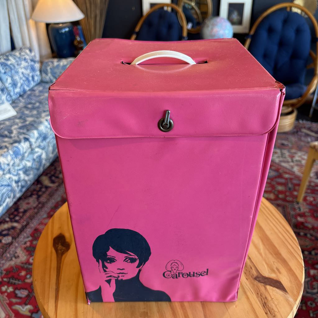 A hot pink wig case box with an image of Twiggy on the front and the word "Carousel" sitting on a table in a room setting.