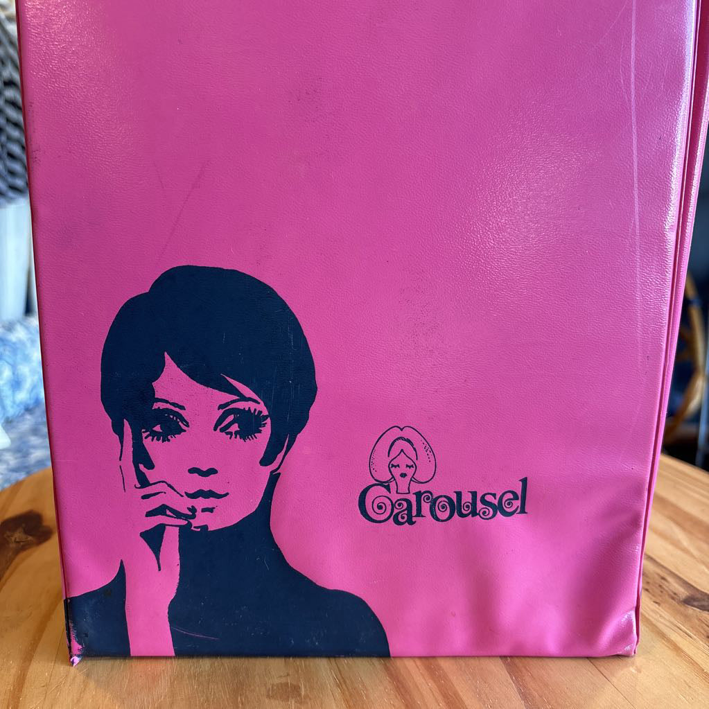 A hot pink wig case box with an image of Twiggy on the front and the word "Carousel" sitting on a table in a room setting.