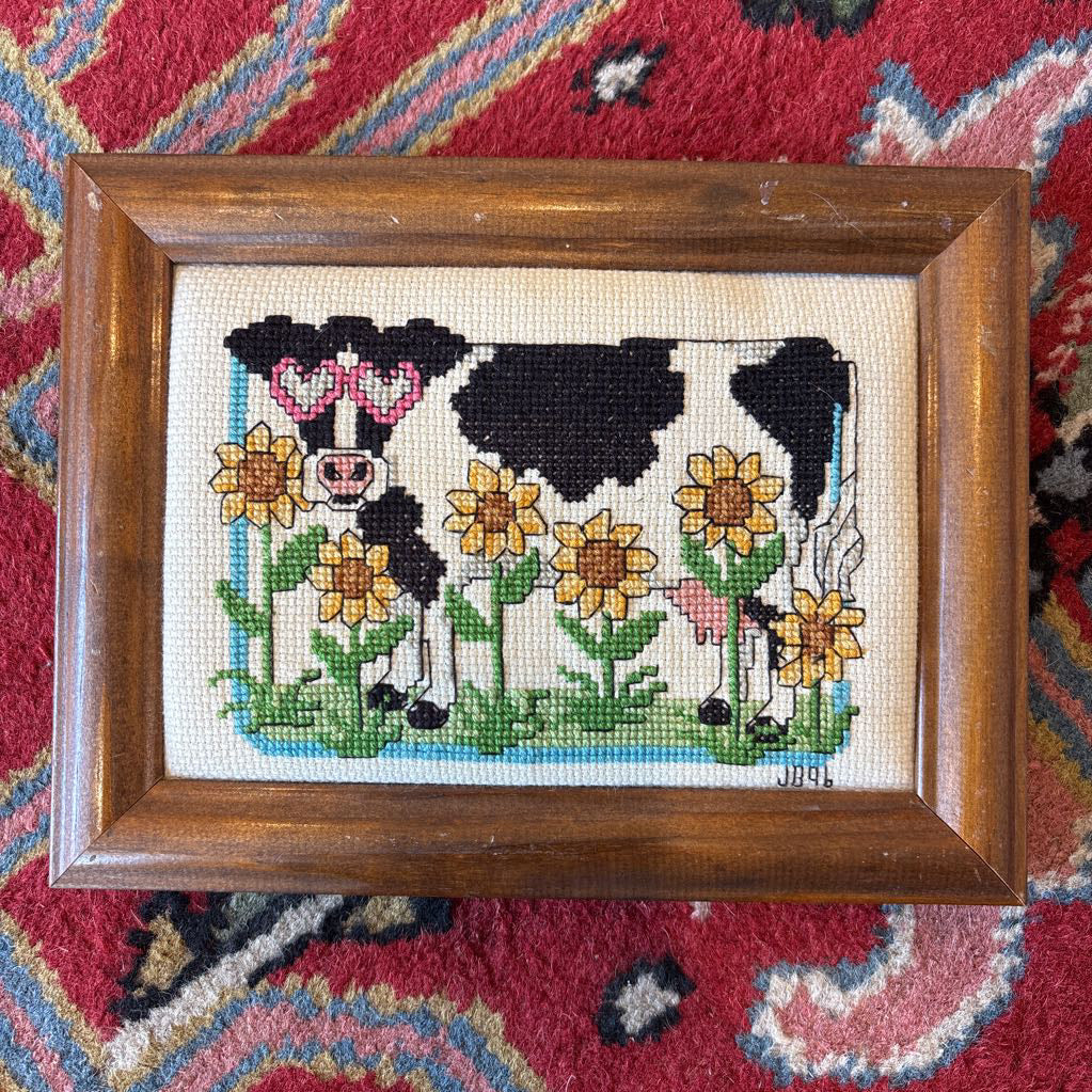 A framed cross stitch artwork of a cow with heart glasses surrounded by sunflowers. This artwork is on a red rug.