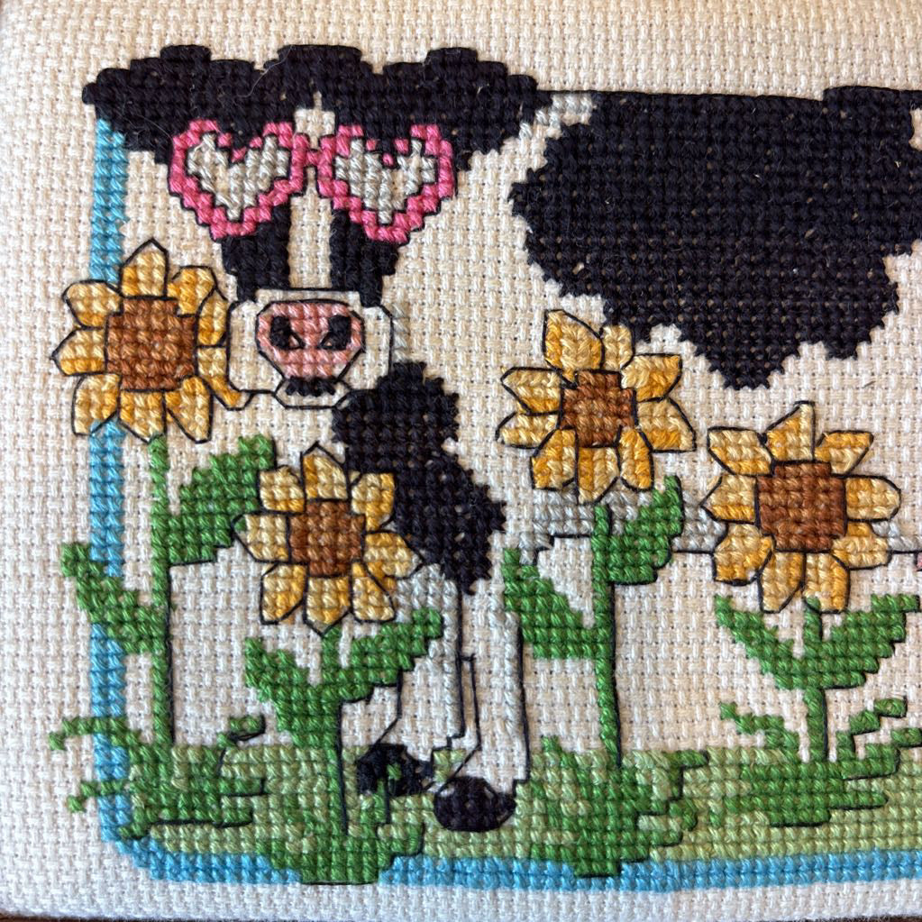 A framed cross stitch artwork of a cow with heart glasses surrounded by sunflowers.