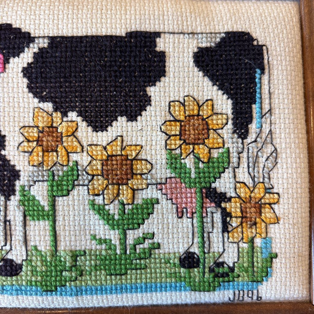 A framed cross stitch artwork of a cow with heart glasses surrounded by sunflowers. This artwork is on a red rug.