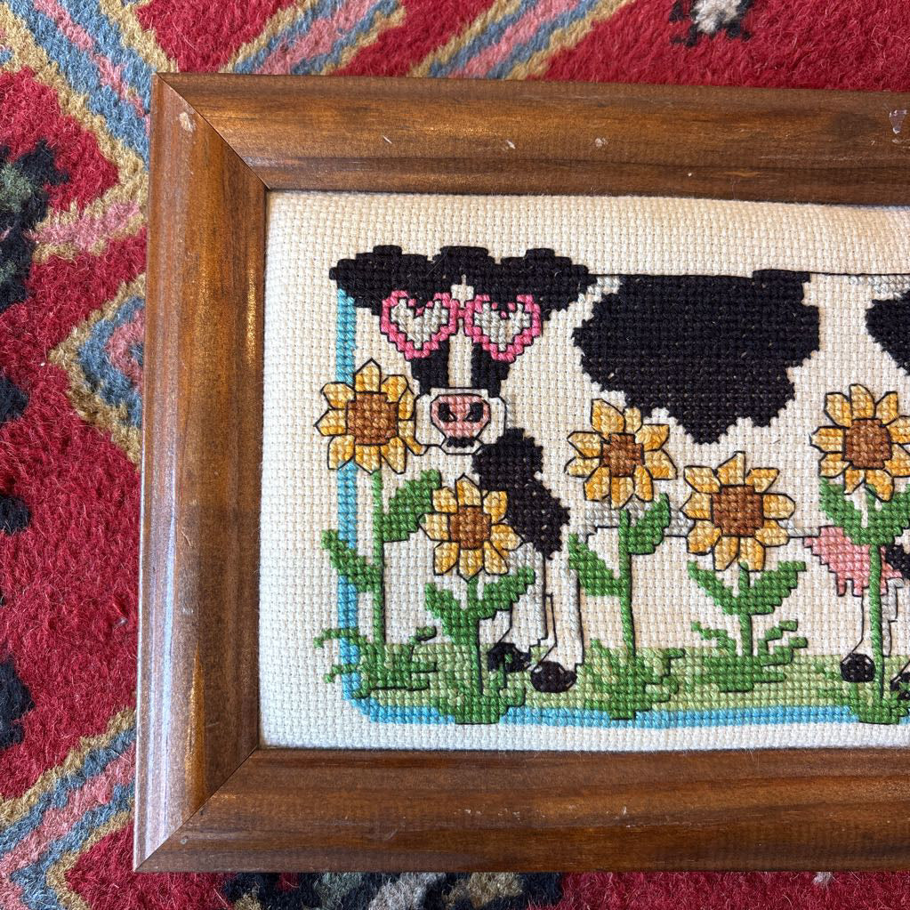 A framed cross stitch artwork of a cow with heart glasses surrounded by sunflowers. This artwork is on a red rug.
