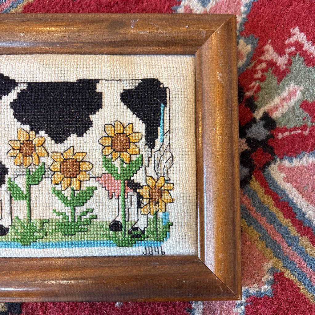 A framed cross stitch artwork of a cow with heart glasses surrounded by sunflowers. This artwork is on a red rug.