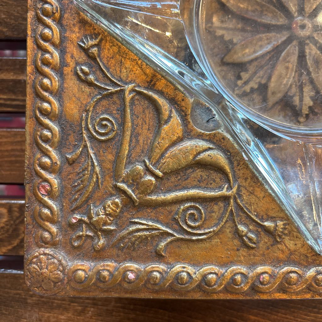 An intricate gold chalkware ashtray with designs of stylized women on the corners and a flower in the center. A glass diamond sits in the center to act as the ashtray. This whole piece sits on a wooden bench.