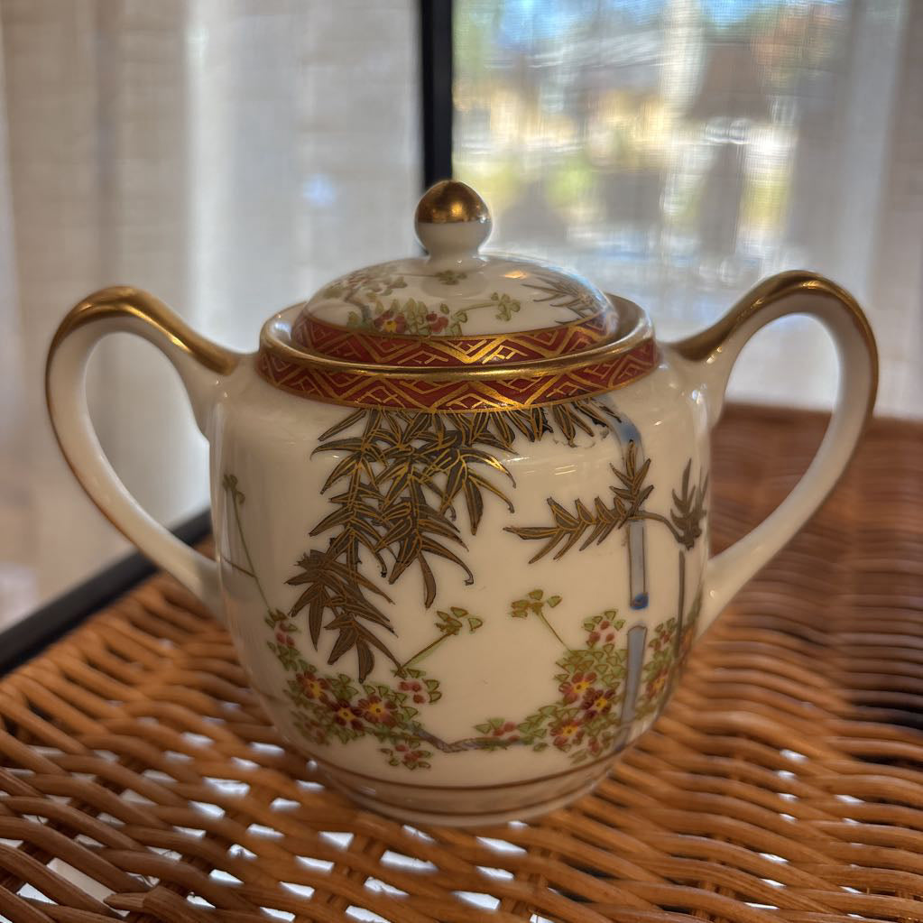 A ceramic lidded sugar bowl with painted floral elements on a rattan self in a room setting.