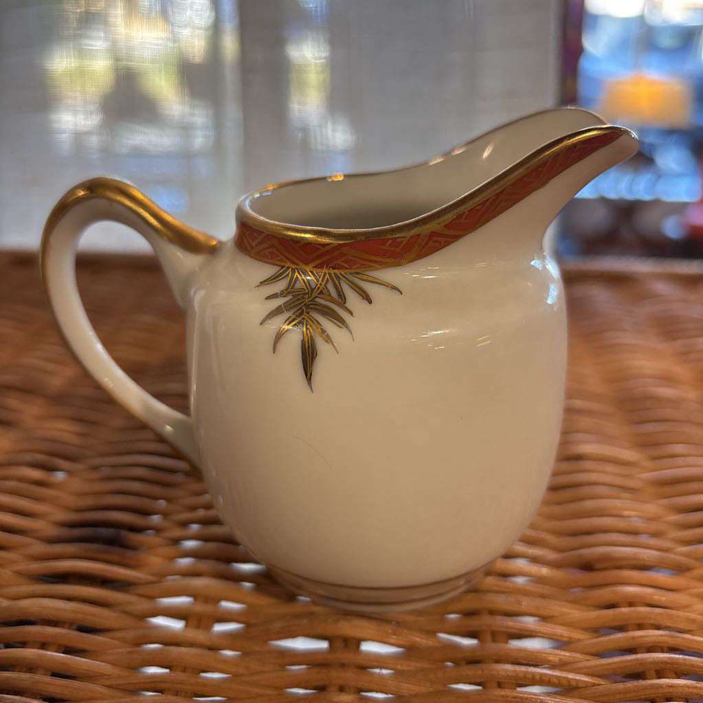 A ceramic creamer with painted floral elements on a rattan self in a room setting.