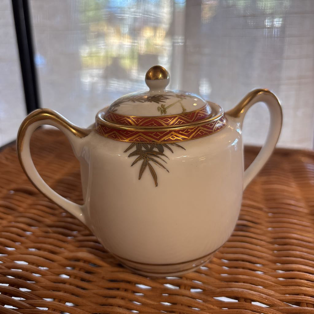 A ceramic lidded sugar bowl with painted floral elements on a rattan self in a room setting.