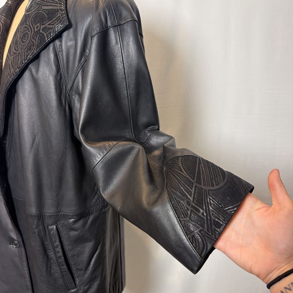 A black leather trench coat with wide shoulders and an abstract design on the lapels. Its on a mannequin in front of a white wall.