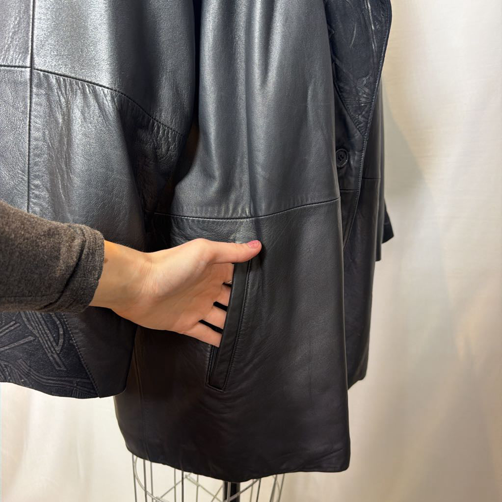A black leather trench coat with wide shoulders and an abstract design on the lapels. Its on a mannequin in front of a white wall.