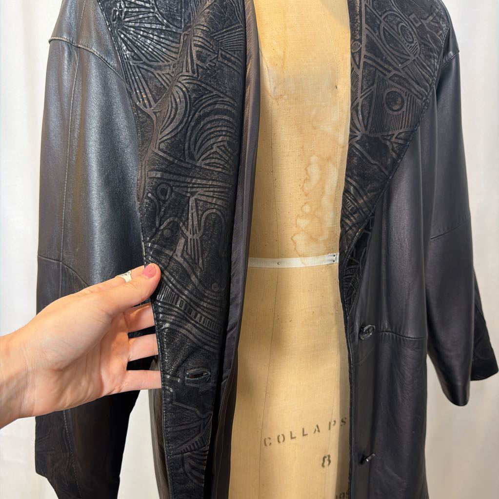 A black leather trench coat with wide shoulders and an abstract design on the lapels. Its on a mannequin in front of a white wall.