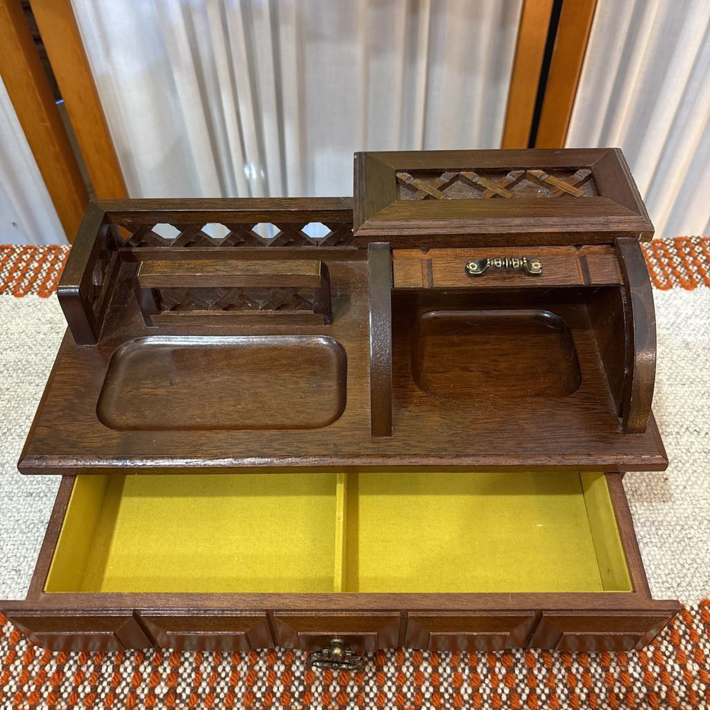 An open wooden jewelry box/valet box on a woven runner in front of a white screen with part of a yellow painting showing.