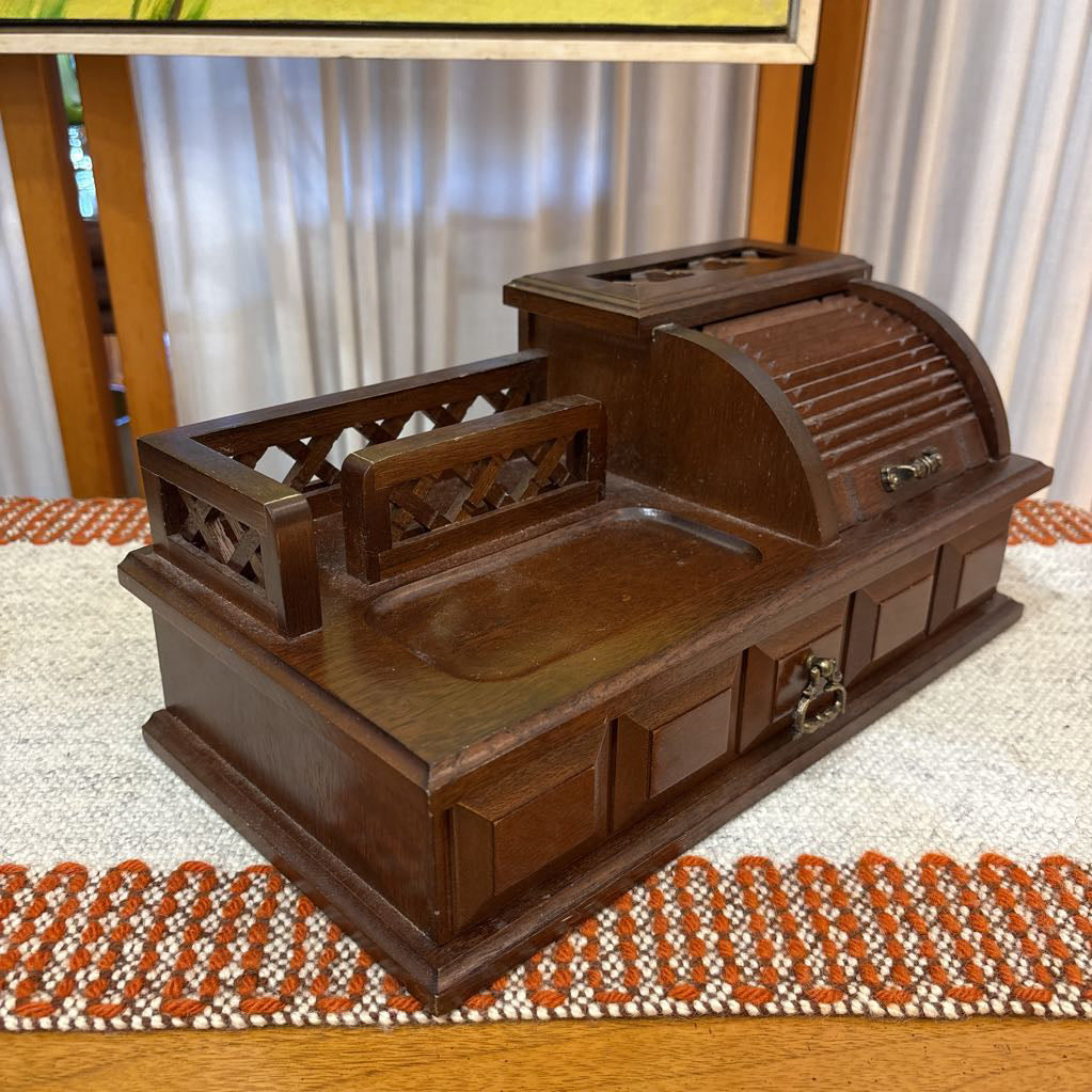 A wooden jewelry box/valet box on a woven runner in front of a white screen with part of a yellow painting showing.