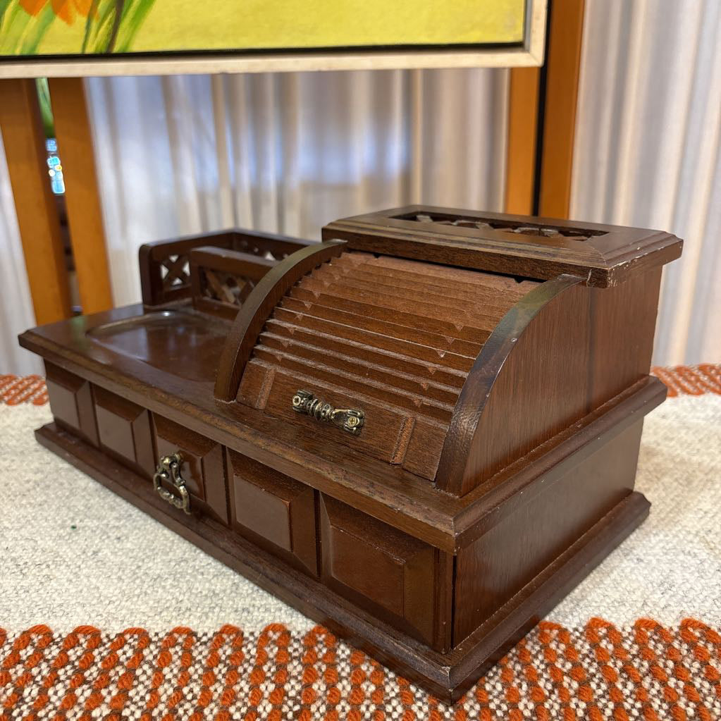A wooden jewelry box/valet box on a woven runner in front of a white screen with part of a yellow painting showing.