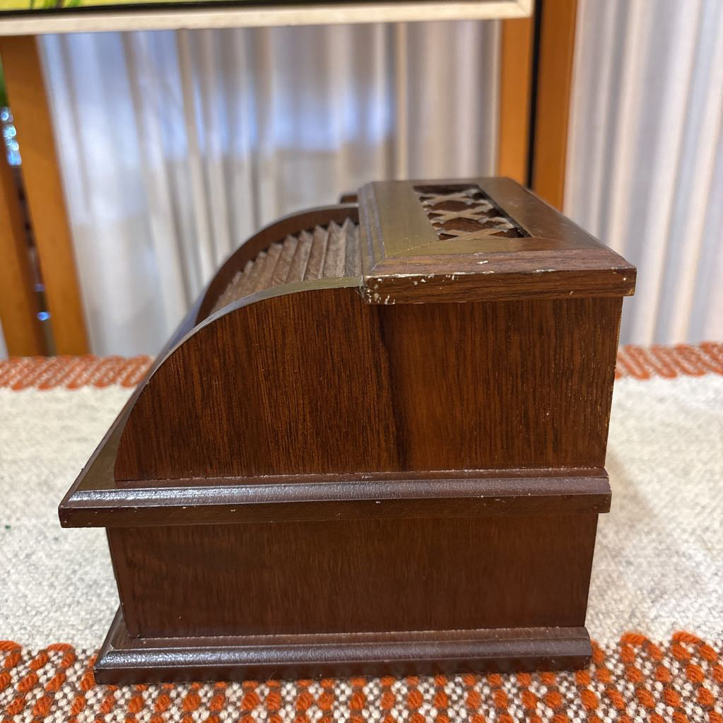 A wooden jewelry box/valet box on a woven runner in front of a white screen with part of a yellow painting showing.