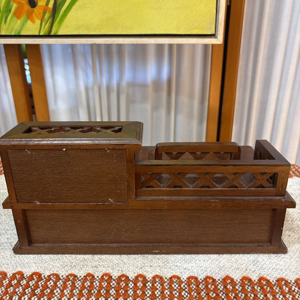 A wooden jewelry box/valet box on a woven runner in front of a white screen with part of a yellow painting showing.