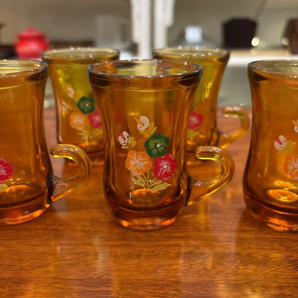 An amber glassware set with a decanter and 5 cordial glasses all with a floral pattern on the front sitting on a wooden table in a store setting.