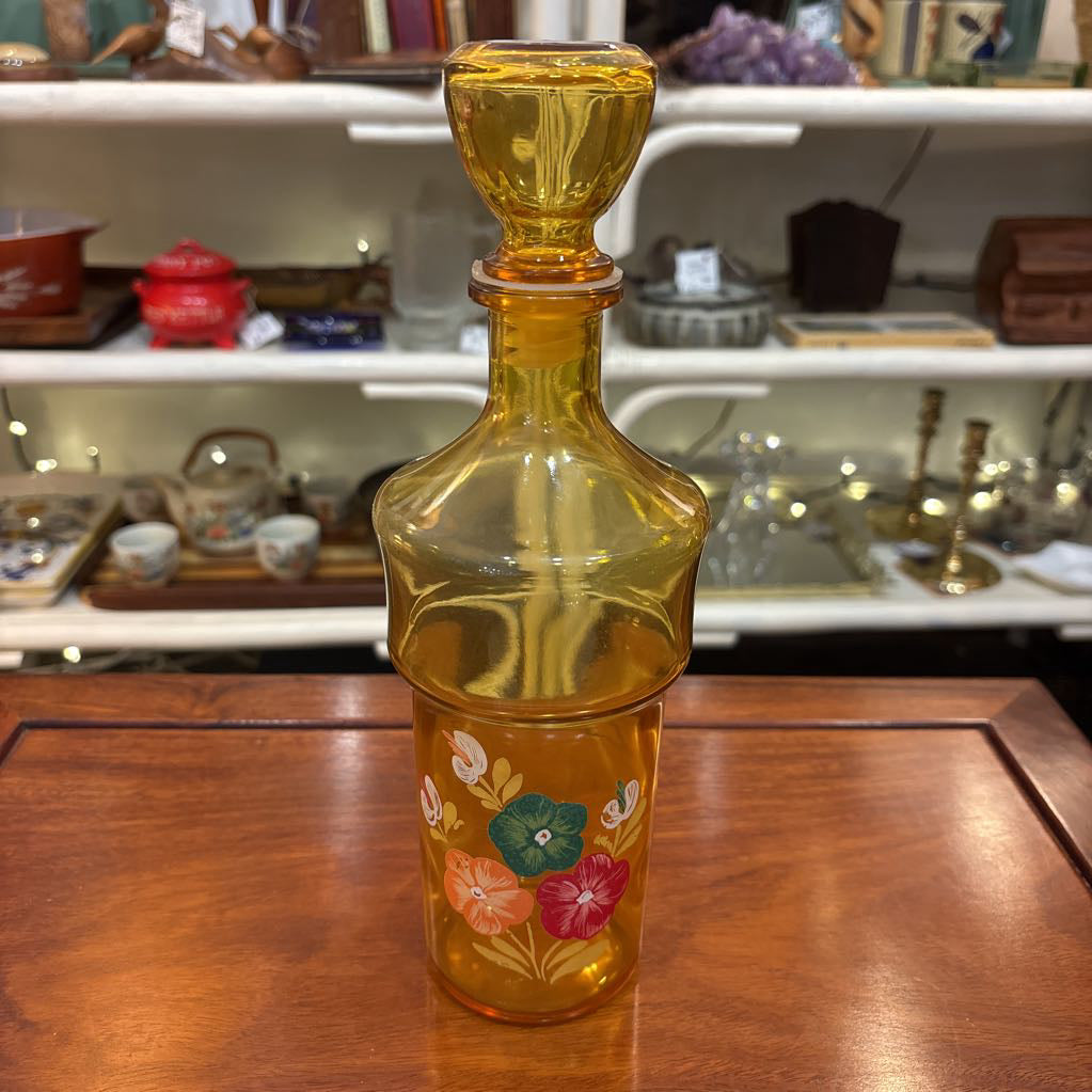An amber glassware set with a decanter and 5 cordial glasses all with a floral pattern on the front sitting on a wooden table in a store setting.