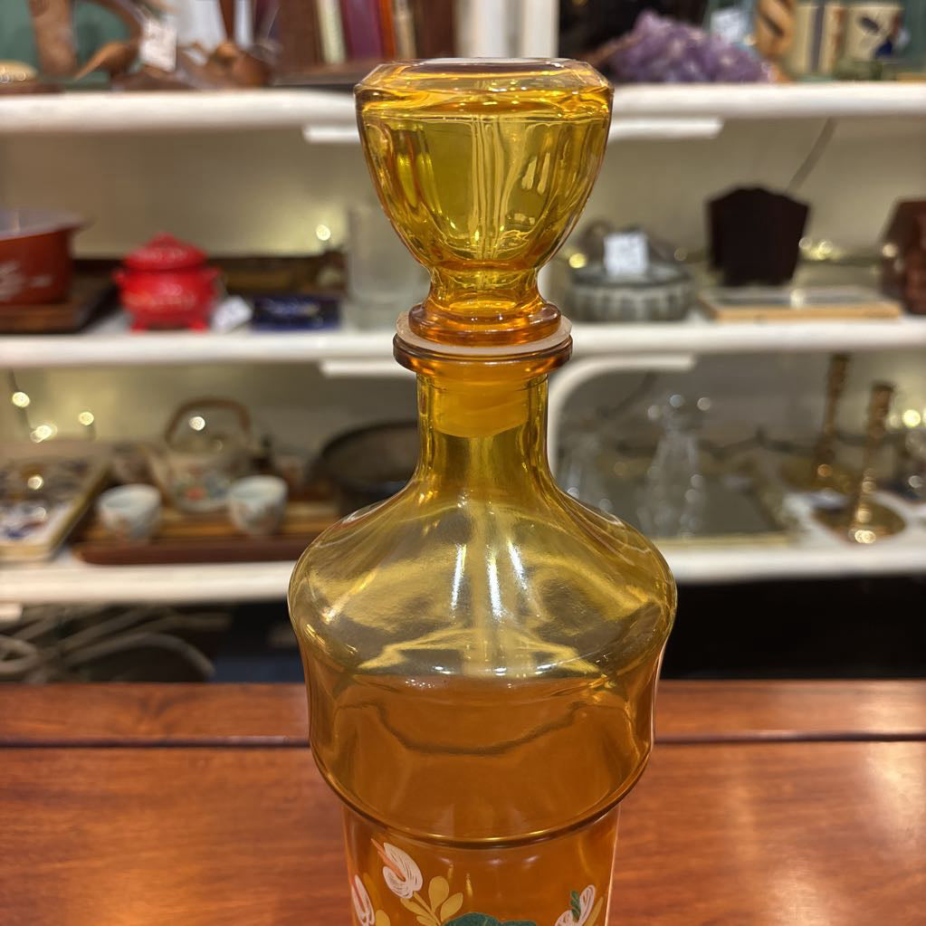 An amber glassware set with a decanter and 5 cordial glasses all with a floral pattern on the front sitting on a wooden table in a store setting.