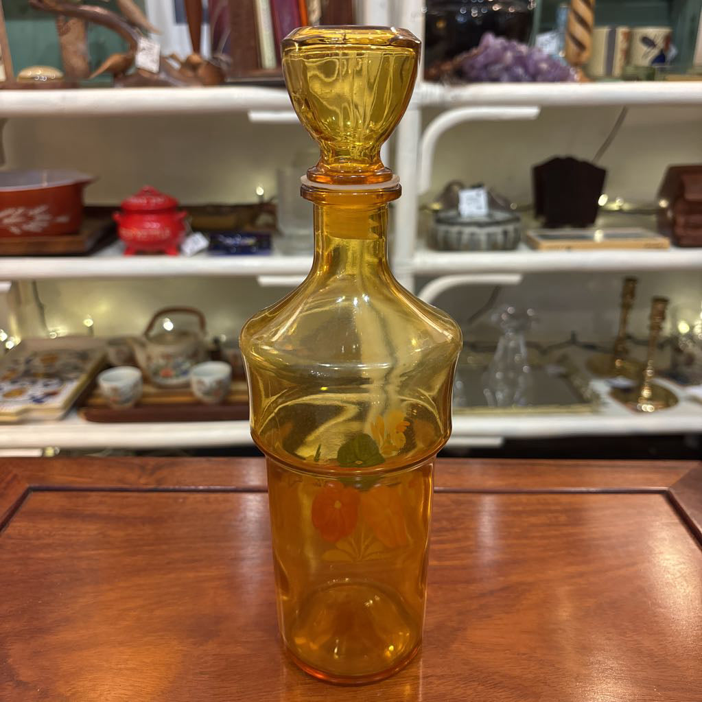 An amber glassware set with a decanter and 5 cordial glasses all with a floral pattern on the front sitting on a wooden table in a store setting.