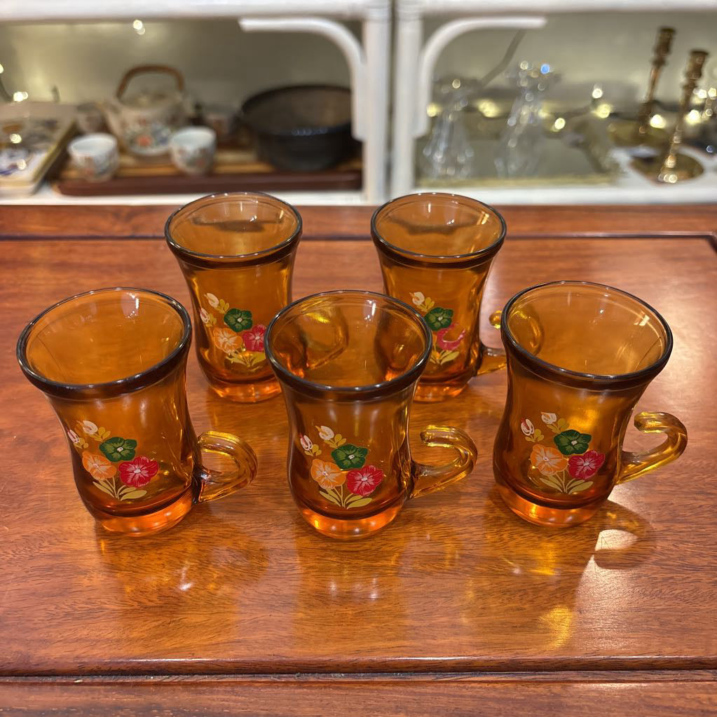 An amber glassware set with a decanter and 5 cordial glasses all with a floral pattern on the front sitting on a wooden table in a store setting.