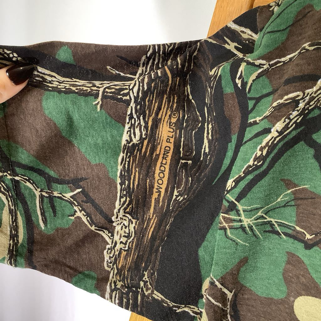 close up of a camo sleeve with the label "Woodland Plus" a hand holding out the sleeve against a white background.