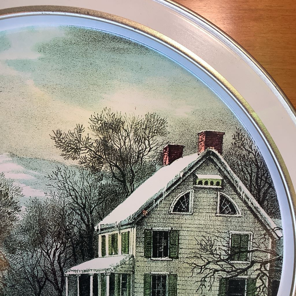 Decorative plate with a winter scene of a house and trees, framed by a metallic border.