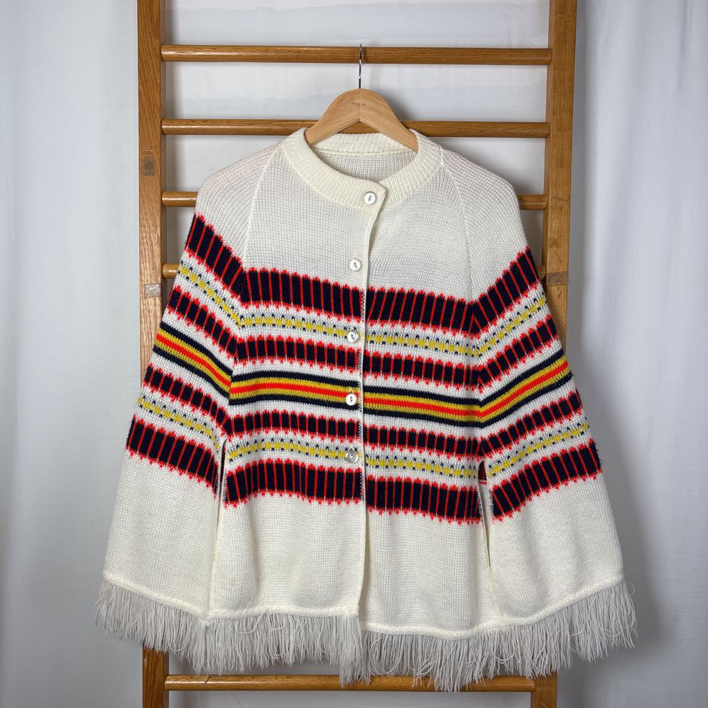 A white striped knitted poncho with a white fringe bottom. Its hanging on a wooden rack. 