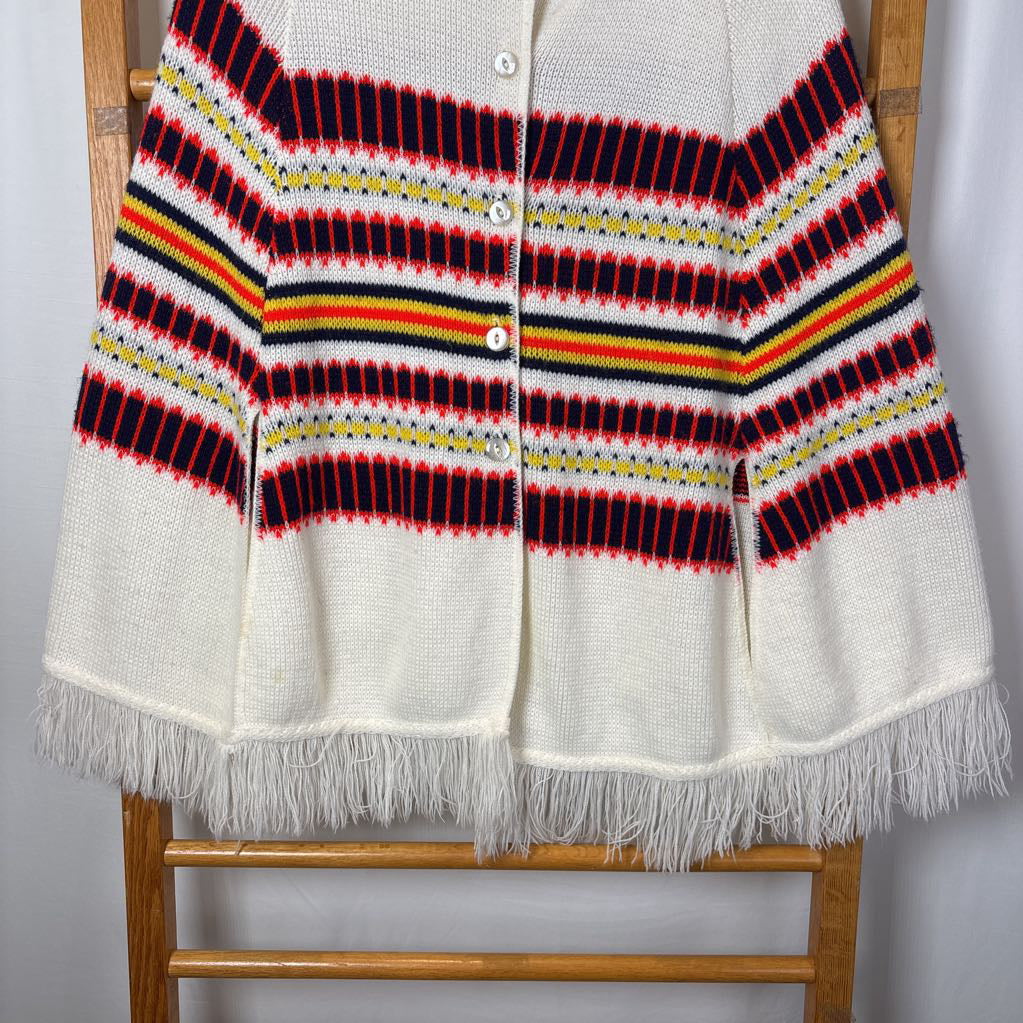A white striped knitted poncho with a white fringe bottom. Its hanging on a wooden rack. 