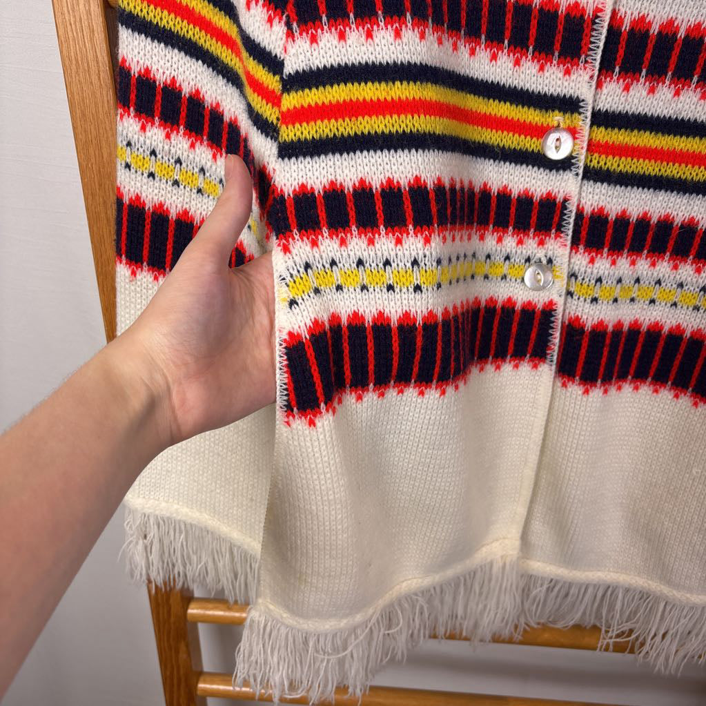 A white striped knitted poncho with a white fringe bottom. Its hanging on a wooden rack. 