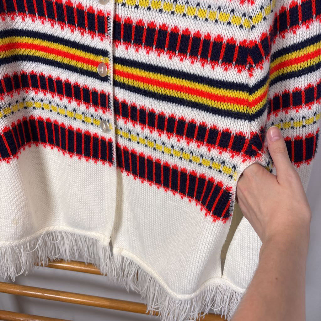 A white striped knitted poncho with a white fringe bottom. Its hanging on a wooden rack. 
