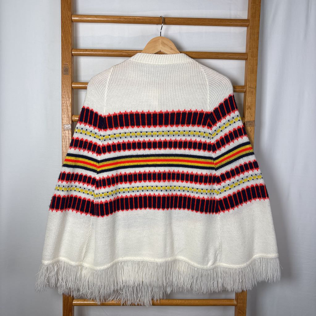 A white striped knitted poncho with a white fringe bottom. Its hanging on a wooden rack. 