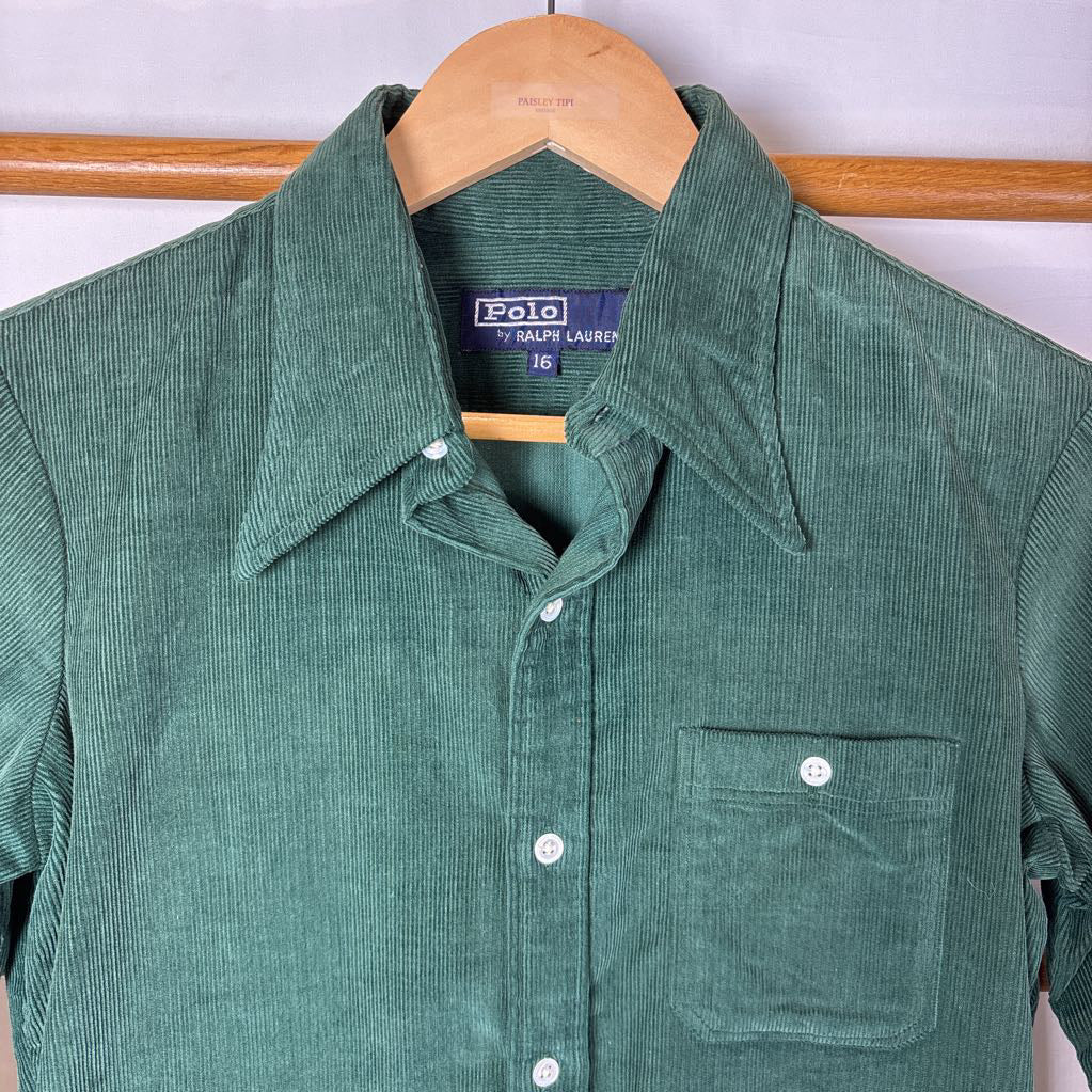A green corduroy button-up long sleeve shirt on a wooden rack against a white wall. 