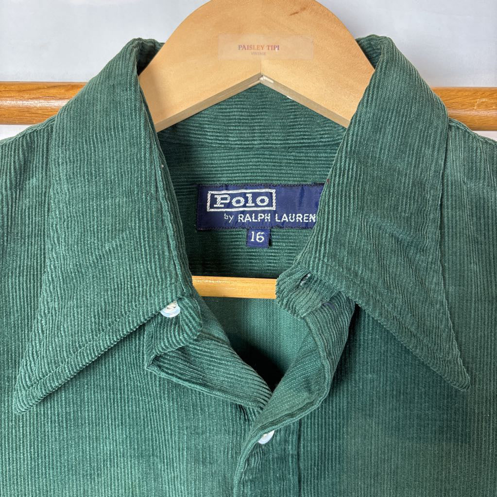 A green corduroy button-up long sleeve shirt on a wooden rack against a white wall. 