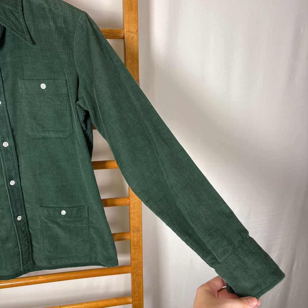 A green corduroy button-up long sleeve shirt on a wooden rack against a white wall. 