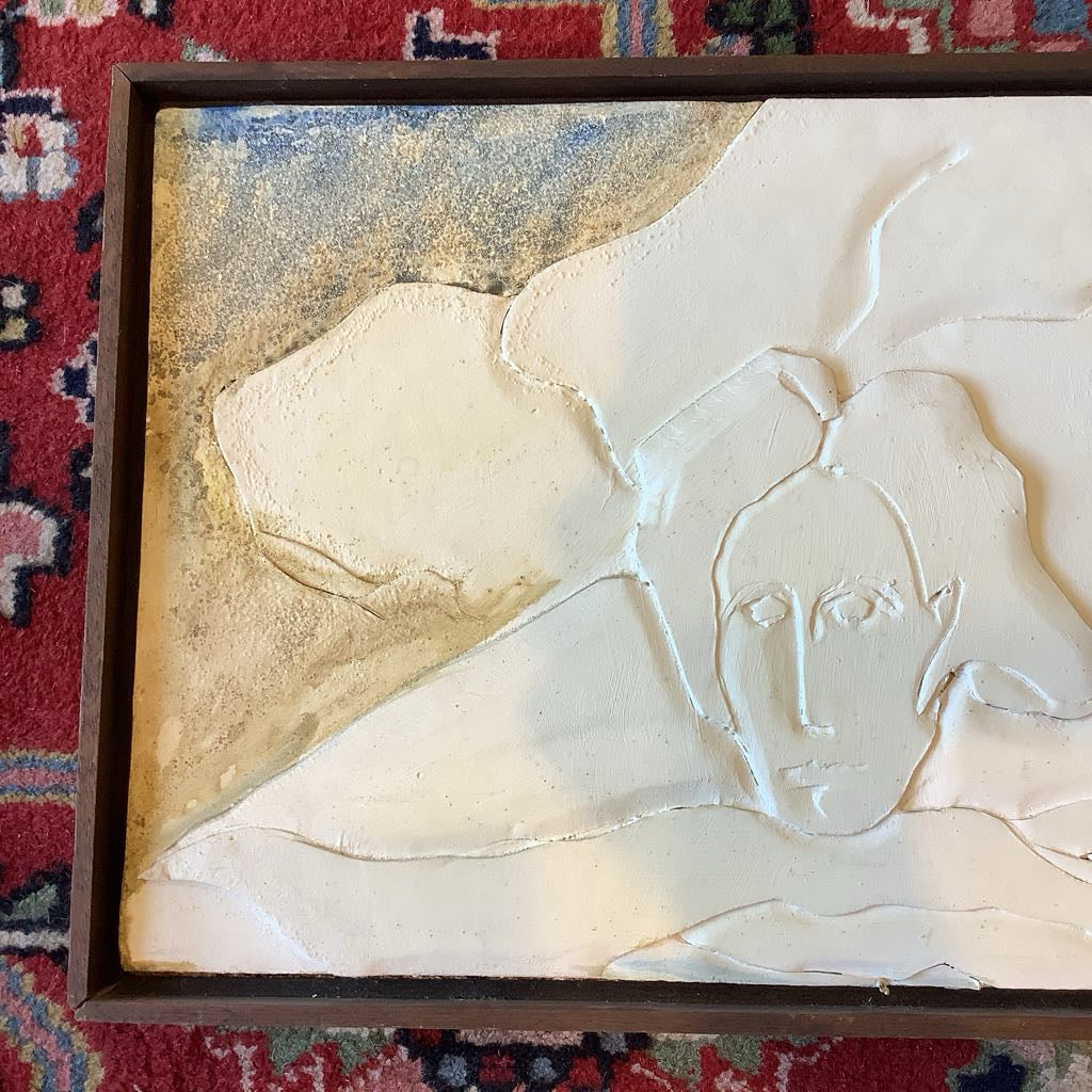 Framed artwork of a stylized face on a textured surface, placed on a patterned rug.