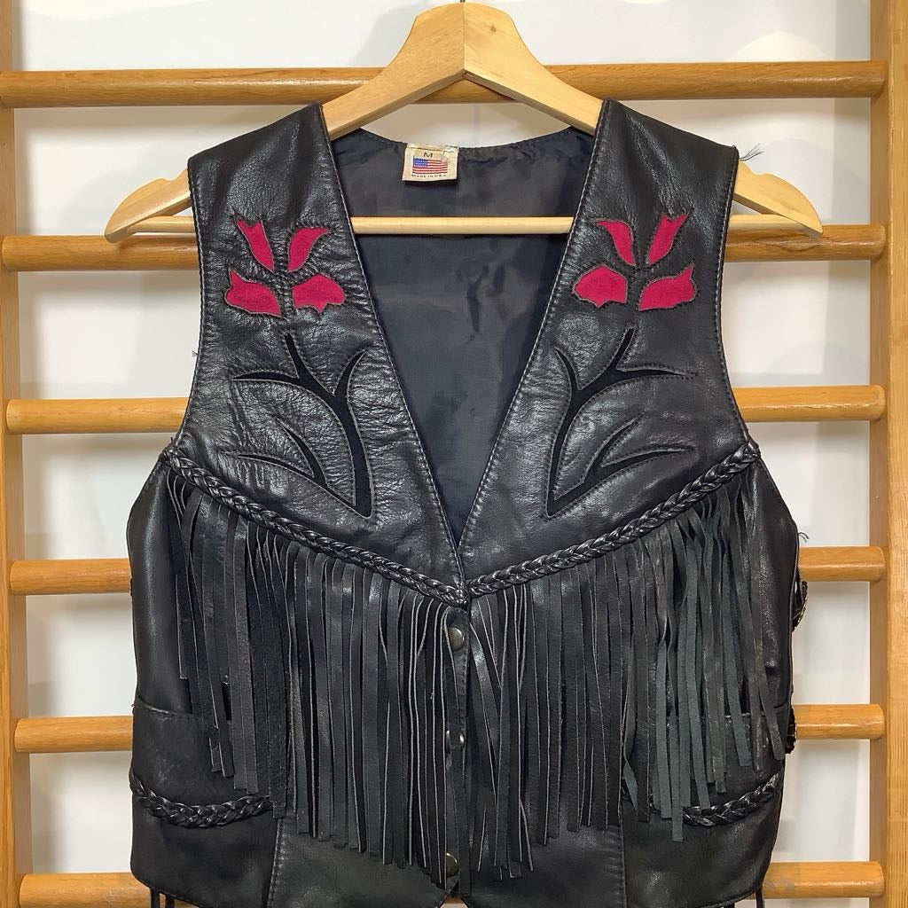 Black leather vest with pink floral designs and fringes on a wooden rack.