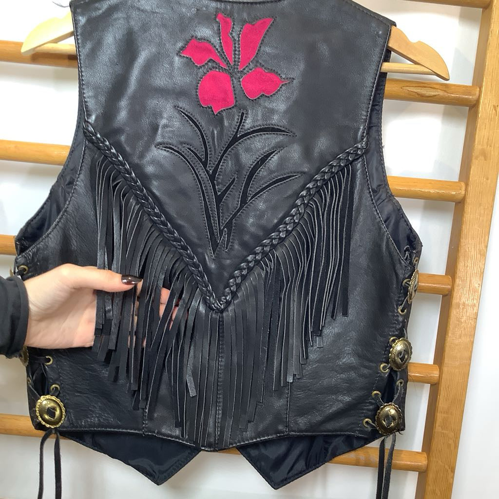 Black leather vest with pink floral design and fringe details on a wooden rack.