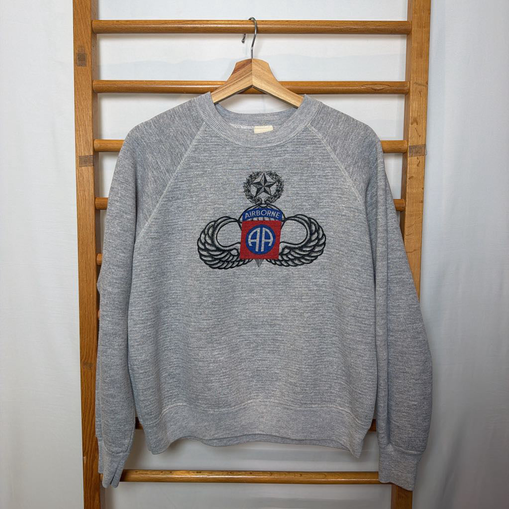 A gray sweatshirt that has a logo and the word "Airborne" on the front hanging on a wooden rack. 