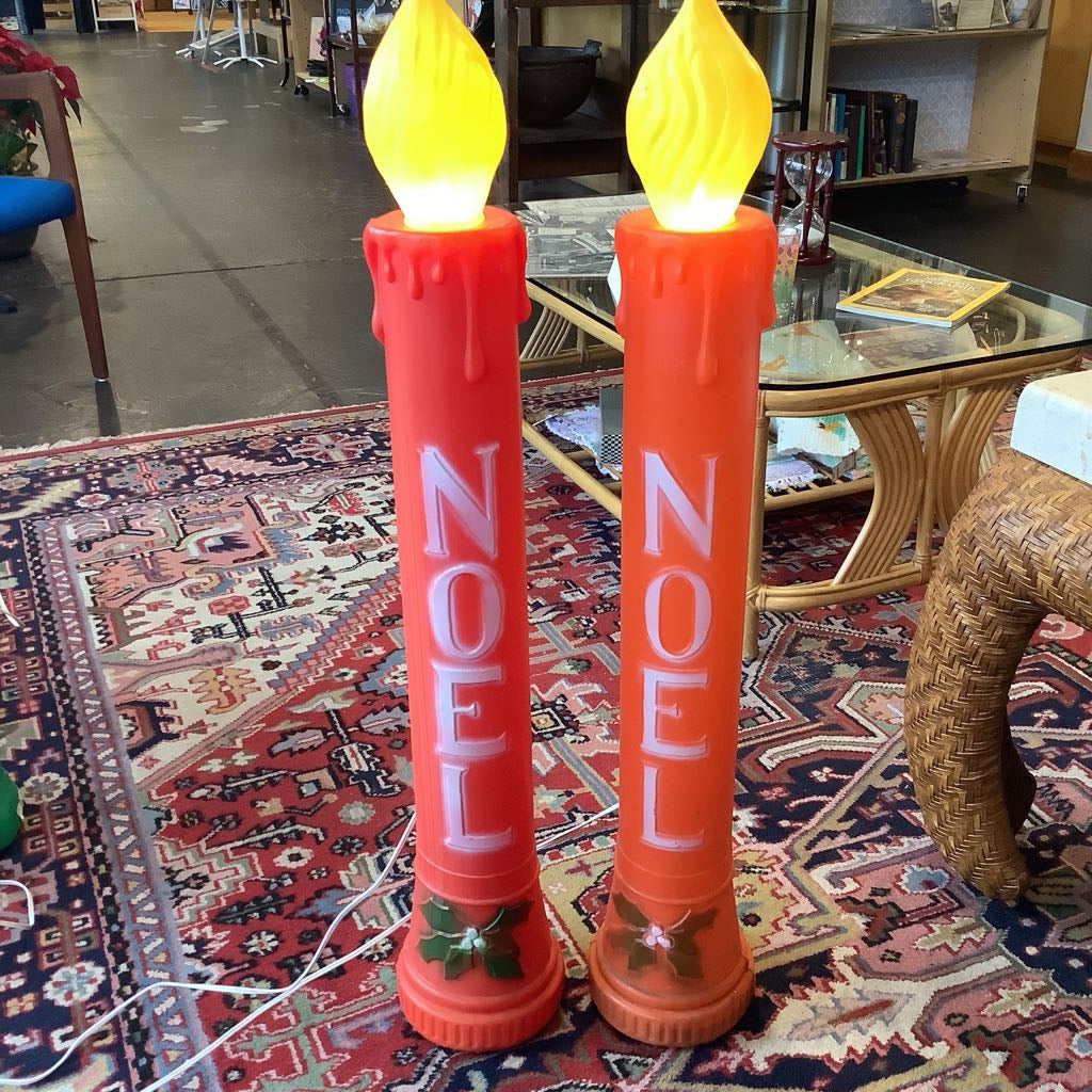 Two orange candles with yellow flames and 'NOEL' text on a patterned rug.
