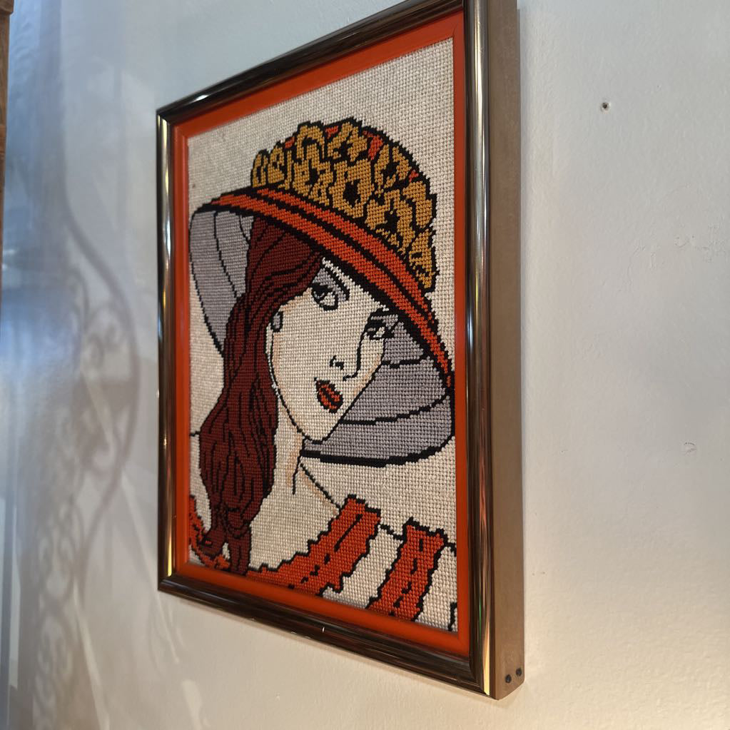 Framed needlepoint of a woman wearing a hat on a white wall