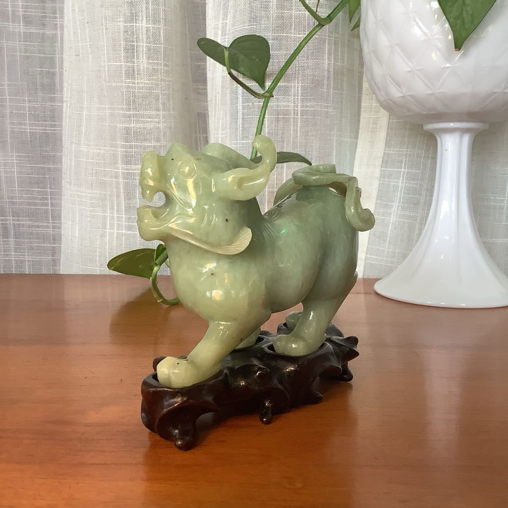 Green jade dragon sculpture on a wooden base with a white vase in the background.