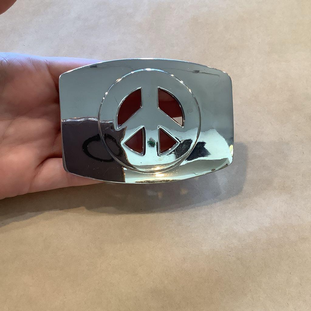 Chrome peace symbol belt buckle held by a hand on a beige background