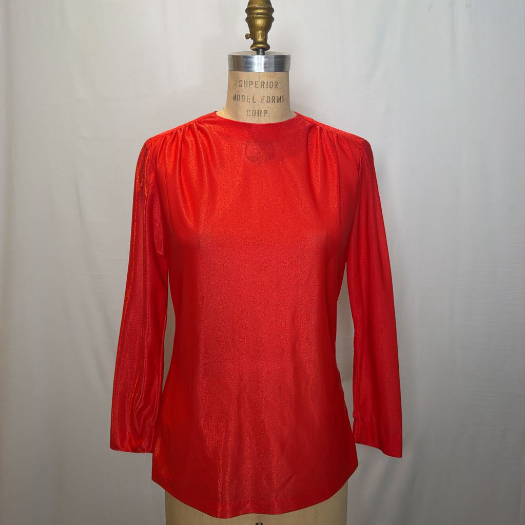 A shimmery red long sleeve blouse on a mannequin in front of a white wall. 