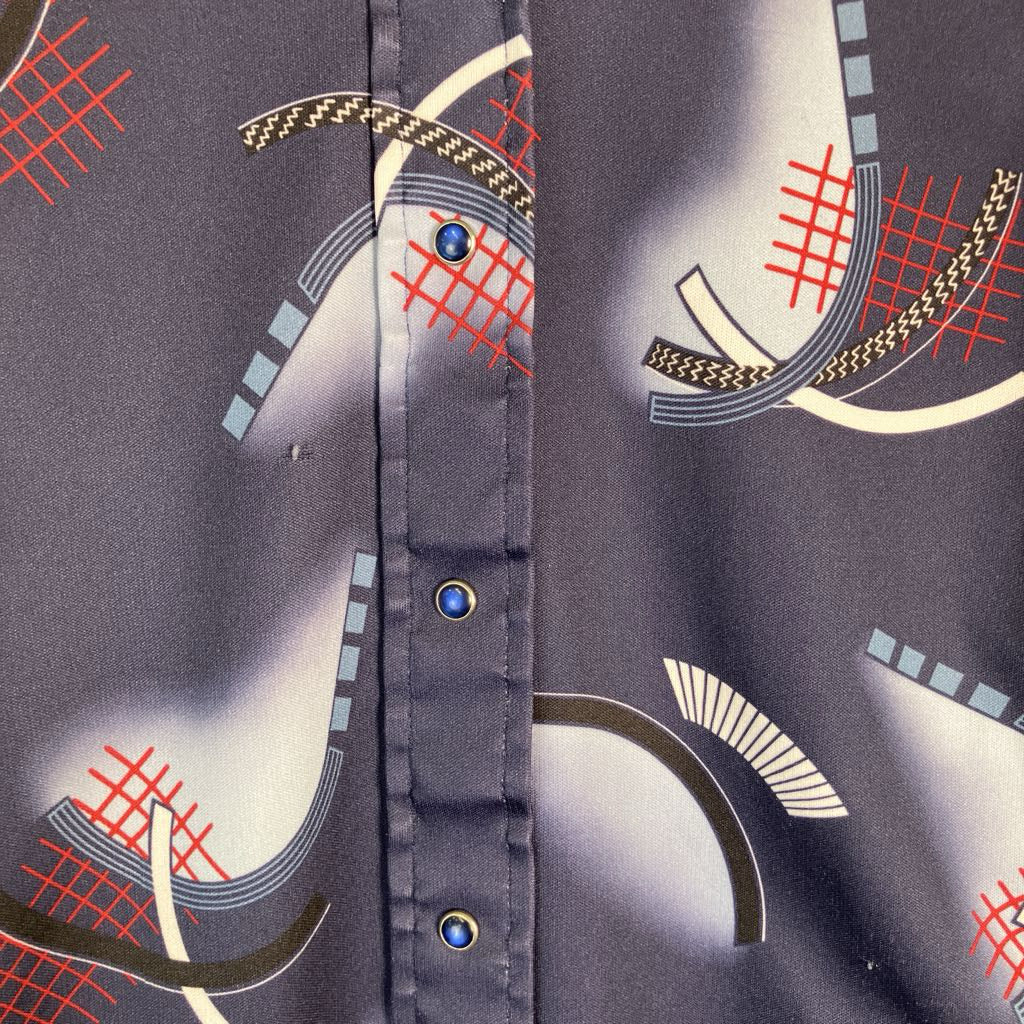 Close-up of a button-front garment fabric with a dark gray base and an abstract print featuring white curved shapes, red grid lines, and small blue button accents.