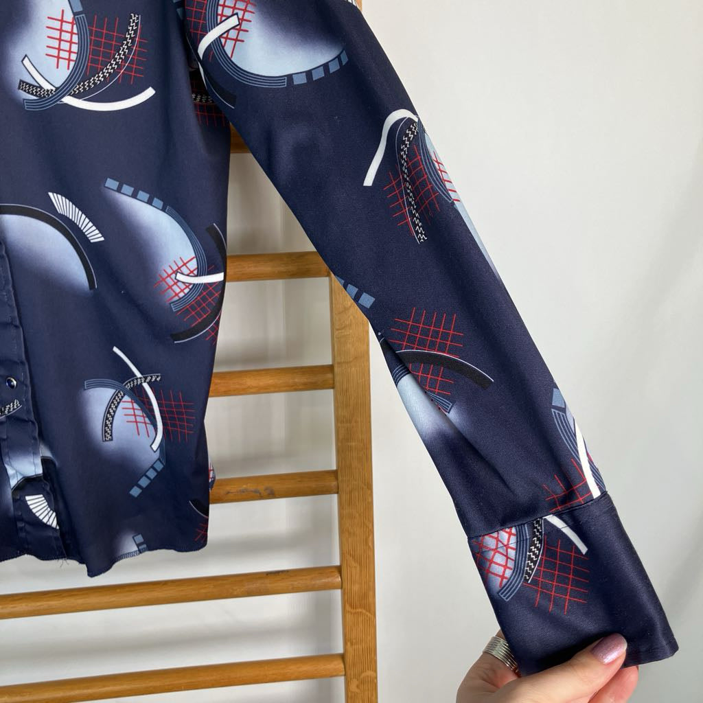 A navy blue long-sleeve top with a tennis-themed graphic print featuring tennis rackets, balls, and grids in red, white, and gray, held up by a wooden ladder with a hand holding the sleeve.