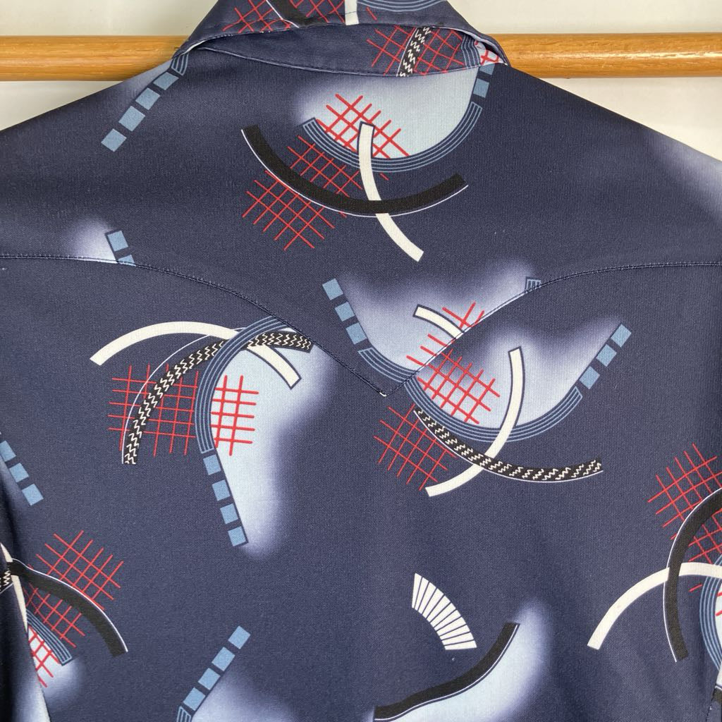 A close-up of a navy blue shirt or jacket featuring a geometric abstract print with white curved lines, red grid patterns, and black accents, hanging on a wooden hanger.