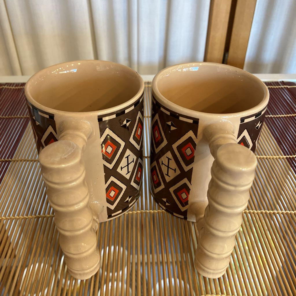 Two ceramic mugs with decorative handles on a woven mat