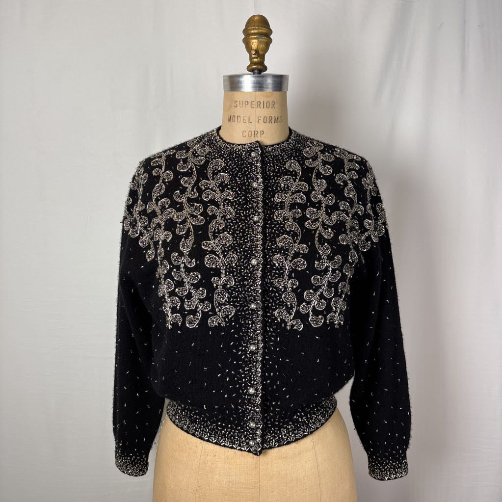 A black button-up sweater with intricate silver bead embroidery on an antique mannequin against a white wall. 